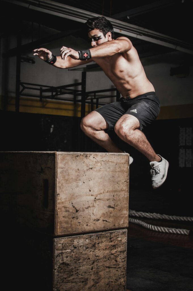 Shirtless man showcasing strength and fitness with a box jump in an indoor gym setting.